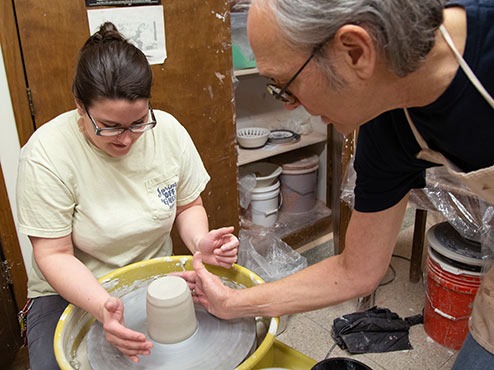 While the student sits at the pottery wheel, the professor begins to instruct them on how to shape their clay.