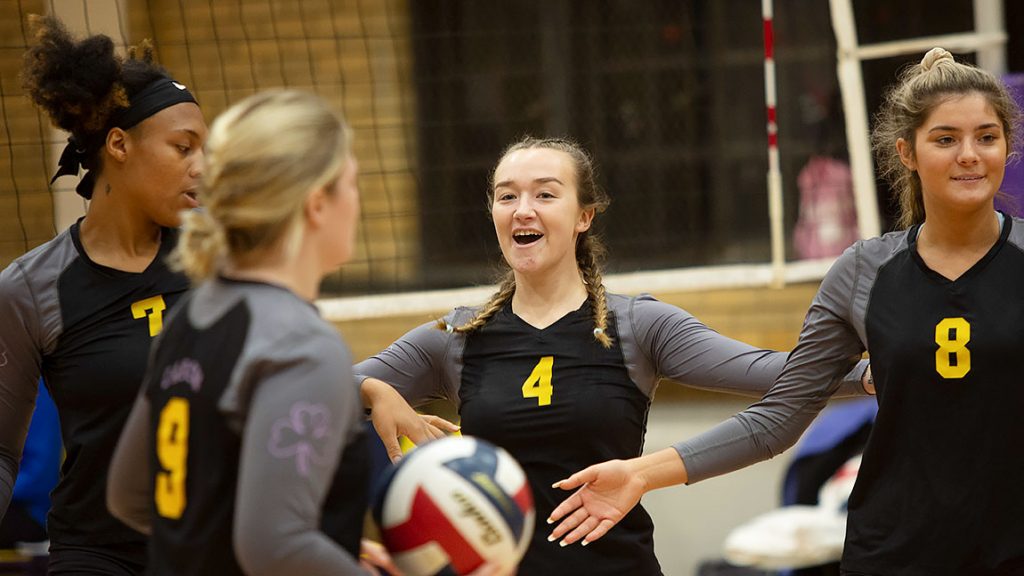 Four students in numbered gray and black shirts get set to play volleyball.