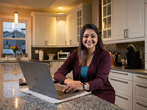 An adult undergraduate sits at her kitchen counter with an open laptop and notepad.