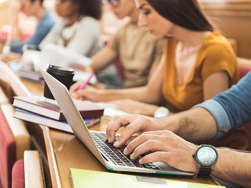 Students sit in class at a row desk. One is typing on a laptop. The others are taking notes.