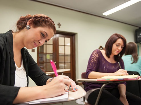 Adult undergraduates write in notebooks while sitting at desks in a classroom.