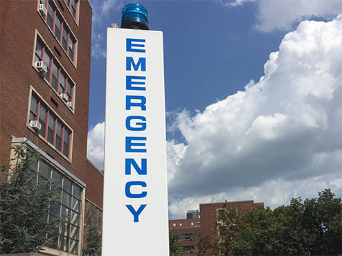A white signpost next to buildings has a blue light on top with “emergency” spelled out vertically in blue letters.