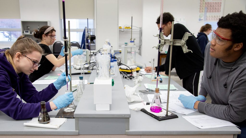 Four students are at a lab table conducting an experiment. Each of them sit at a station with notes and a beaker.