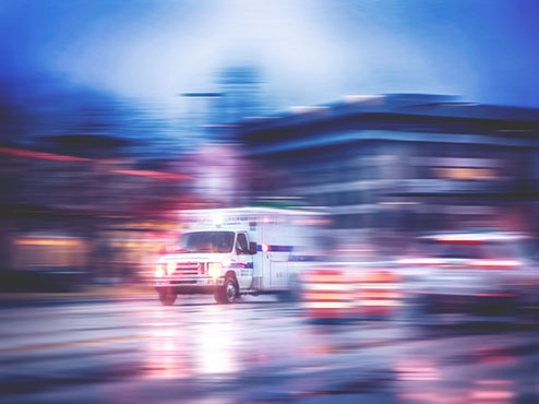 An ambulance rushes down a city street with traffic and buildings blurred out.
