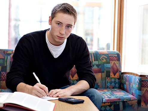 A student sitting on a couch looks up from his work at a table with a book and electronic device in front of him.