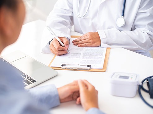 A health services professional in a white coat, sitting across from another person, writes on a form.