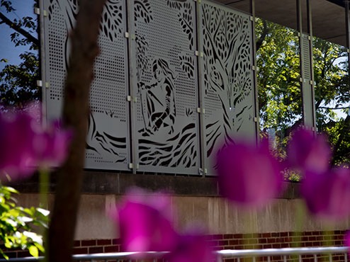 A covered outdoor walkway on campus has artwork on one side and blooming flowers on the other.