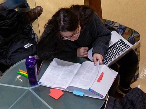 An honors program student reads from a large book while sitting at a small table with an open computer on her lap.