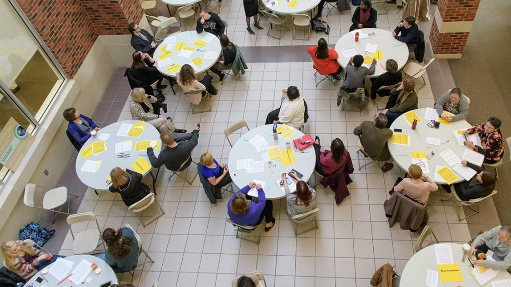 View from above an event shows people sitting at round tables with yellow and white pieces of paper.
