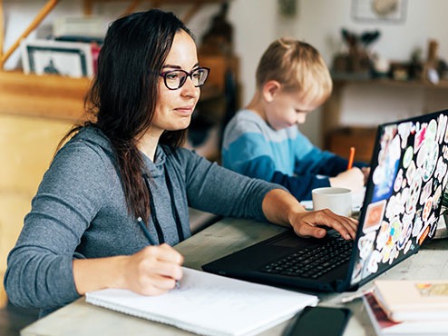 An adult undergraduate student writes in a notebook while reading a laptop screen at home with a young boy beside her.