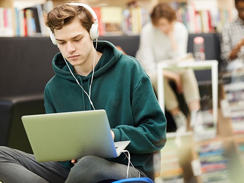 A male student wearing headphones types on a laptop while sitting on the floor. 