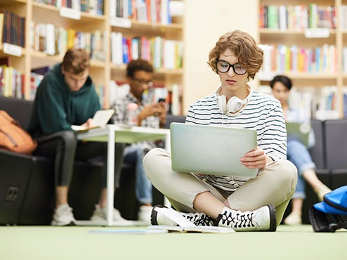 A student sits on the floor with a laptop while others sit around her in a room with bookshelves.