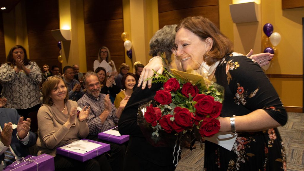 A woman holding red roses hugs another woman at a service awards celebration in front of a small, clapping audience.