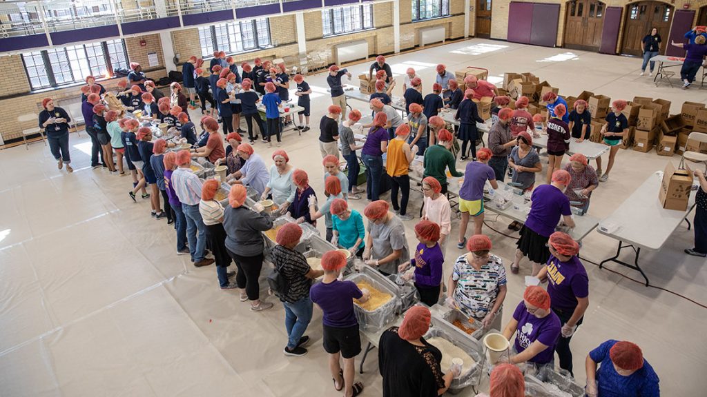 Nearly 100 people wearing orange hairnets stand at tables packing food supplies for Mercy Service in Action.