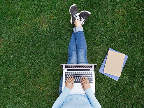 A student types on a laptop while sitting in the grass with books to the side.