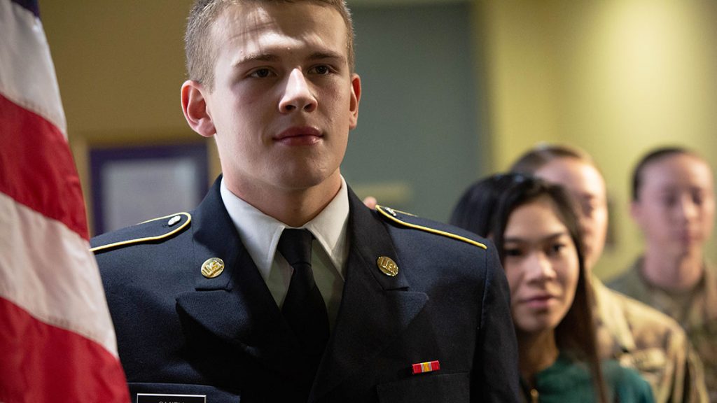 An ROTC student in dress uniform stands next to the U.S. flag and in front of other students at an ROTC recognition event.