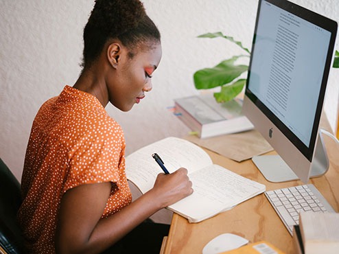 A female student writes in a notebook at a desk in front of a computer.