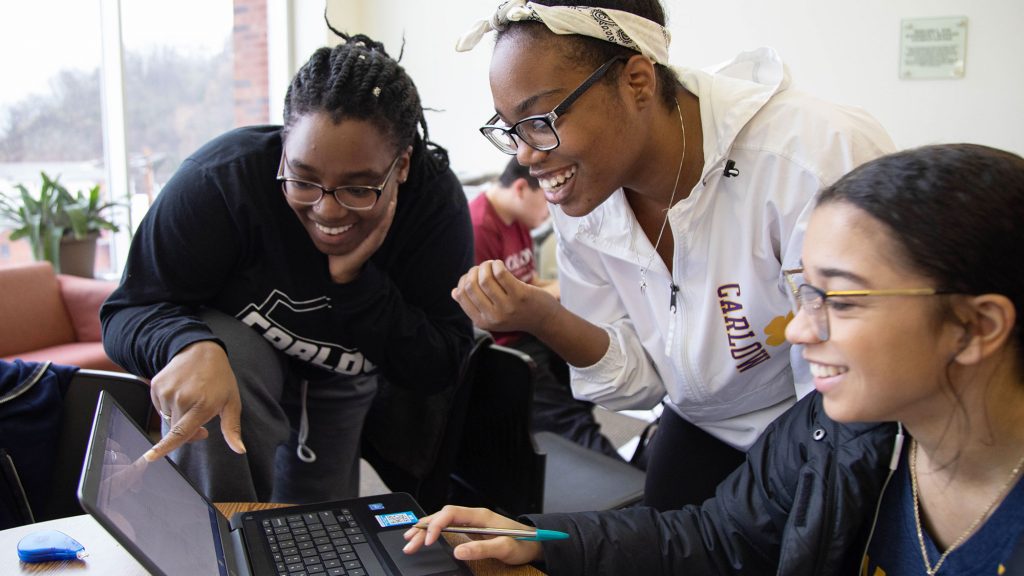 Three students look at a computer screen. One is pointing. They are all smiling.