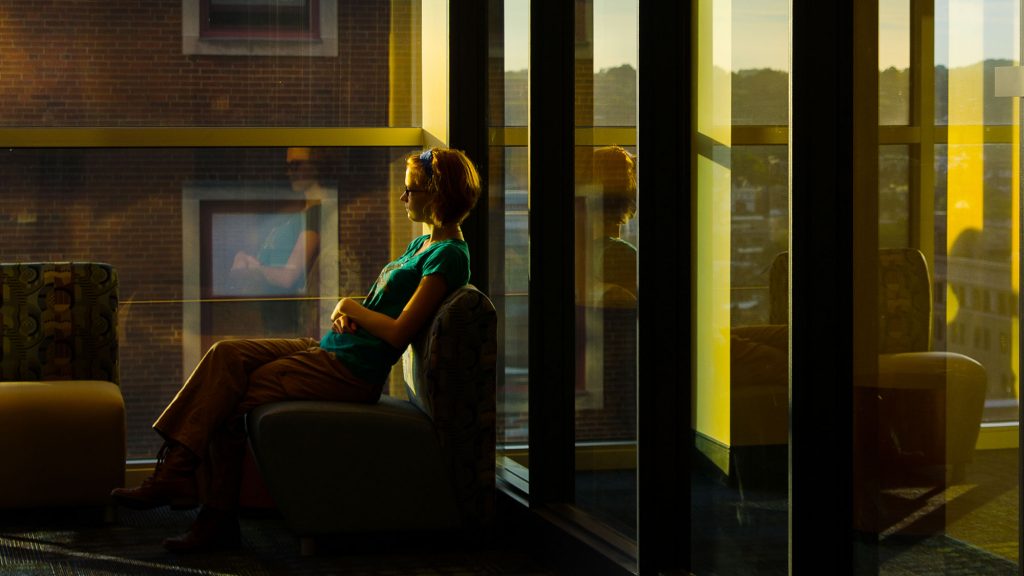 A student gazes out of a window at sunset. The reflection show in the glass behind her.