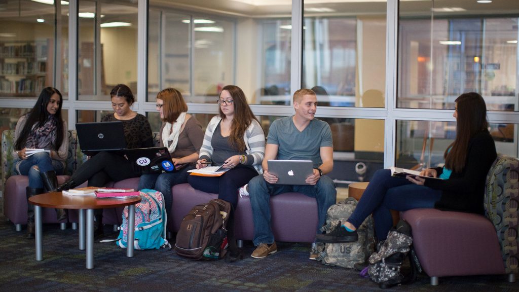 A group of college students sit outside the library. Some are on laptops and some are reading books.