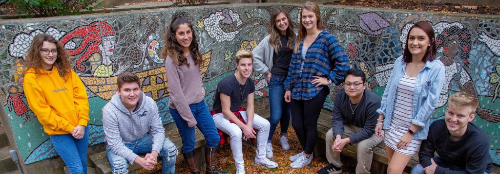 A group of nine students gather on Carlow's campus. Some are standing and some art sitting. Behind them is a colorful mosaic.
