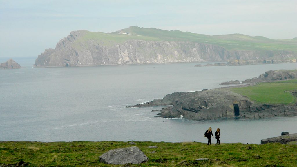 Two people are walking along a cliff and body of water.