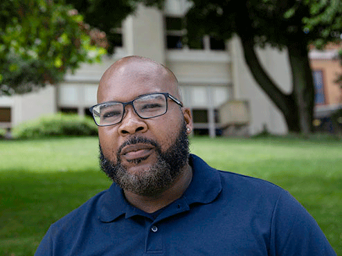 Terrel Williams, a bearded African American man, stands in the shade in front of a field of grass.