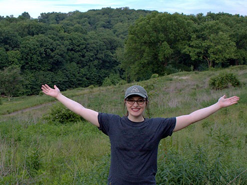 A female student standing with her arms spread in a overgrown field.