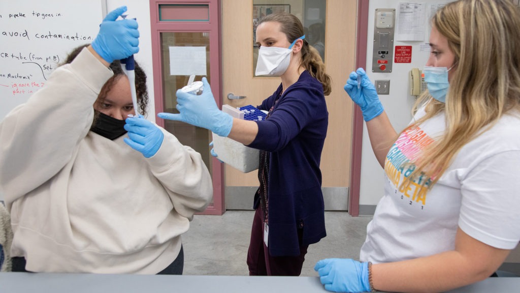 Three students, in blue gloves, stand together conducting an experiment in class at Carlow University.