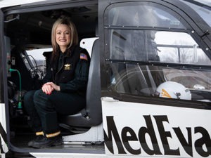 Trista Weiss, wearing a black jacket and black pants, sits in the doorway of a stationary medical helicopter.