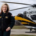 Trista Weiss stands in a black sweater on a helicopter landing pad. A white, black and gold helicopter rests in the background.
