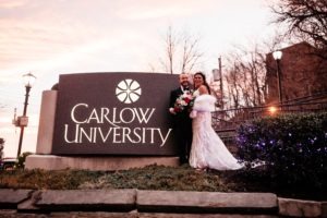 A married couple stands next to the Carlow University sign on campus.