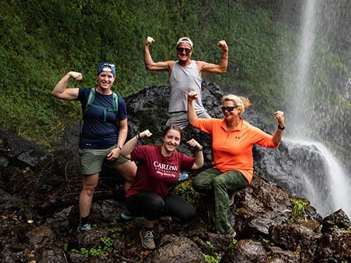 Three women and one man stand on top of a rock flexing their muscles. A waterfall is behind them.