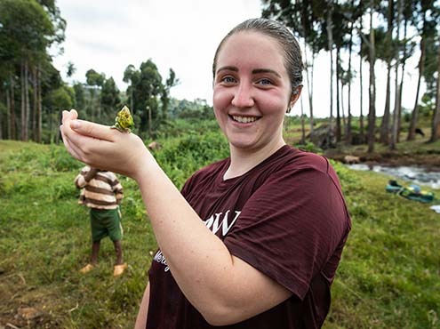 A Carlow student holds a Ugandan reptile in a field near a river.