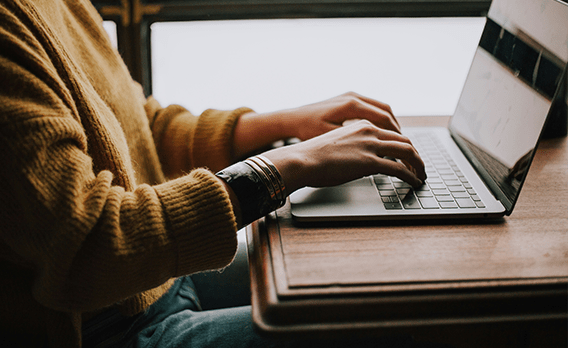 A student in a mustard sweater typing on a laptop at a wooden table