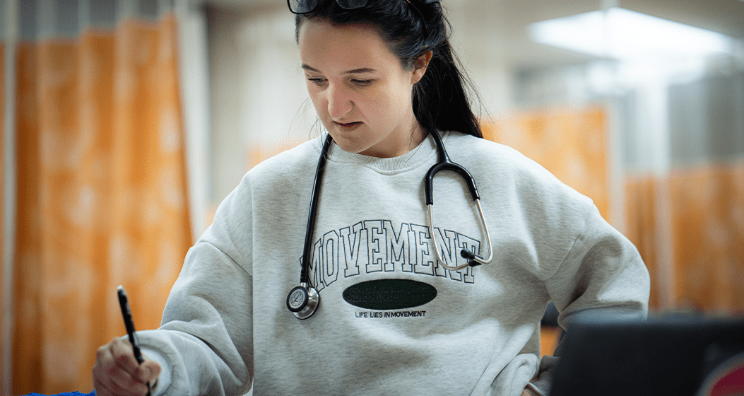 A Carlow University nursing student wearing a stethoscope taking notes in a clinical simulation lab
